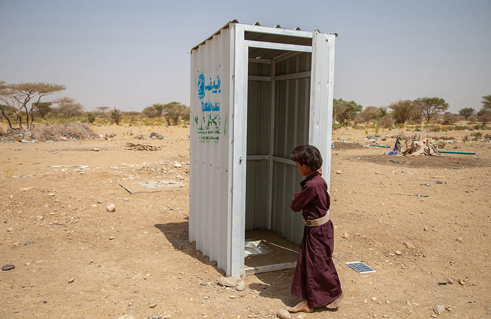 A child stands at the entrance to a latrine made from sheet metal. © UNICEF/UN0495225/Al-Mass